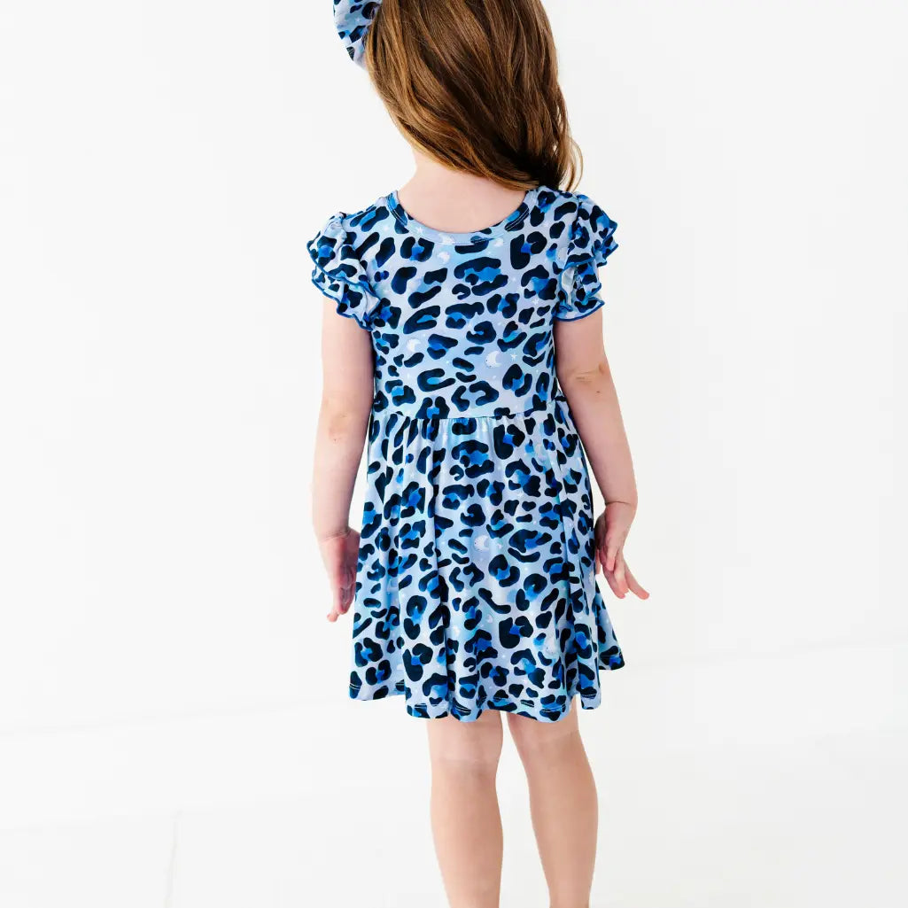Young girl seen from behind wearing a blue leopard-print ruffle-sleeve dress and a matching bow headband, on a white background