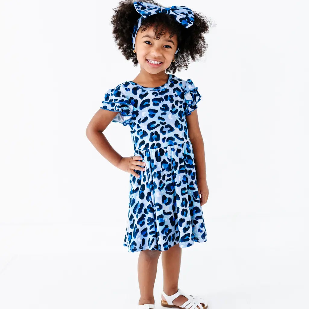 Smiling young girl standing with a hand on her hip, wearing a blue leopard-print ruffle-sleeve dress, a matching bow headband, and white sandals, on a white background