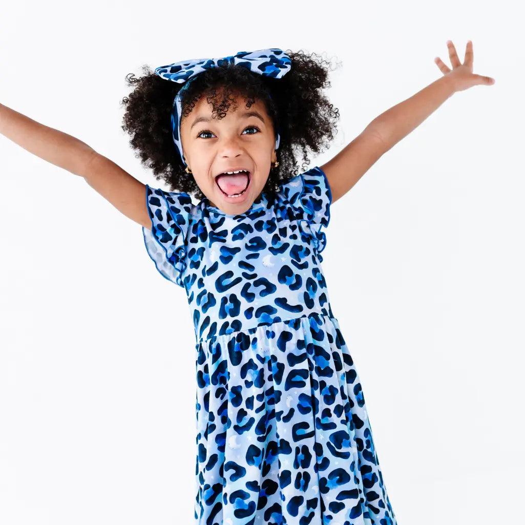 Excited young girl with her arms raised, wearing a blue leopard-print ruffle-sleeve dress and a matching bow headband, on a white background