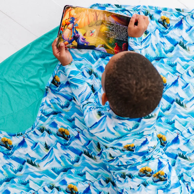 Child wearing a mountain-patterned outfit lying on a matching blanket, reading a book.