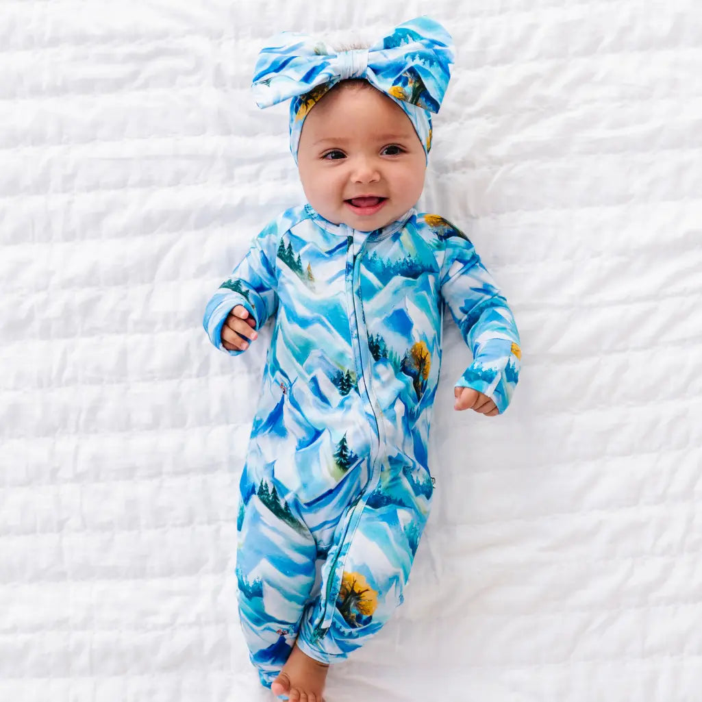 Baby wearing a blue and white patterned onesie and headband on a white textured surface