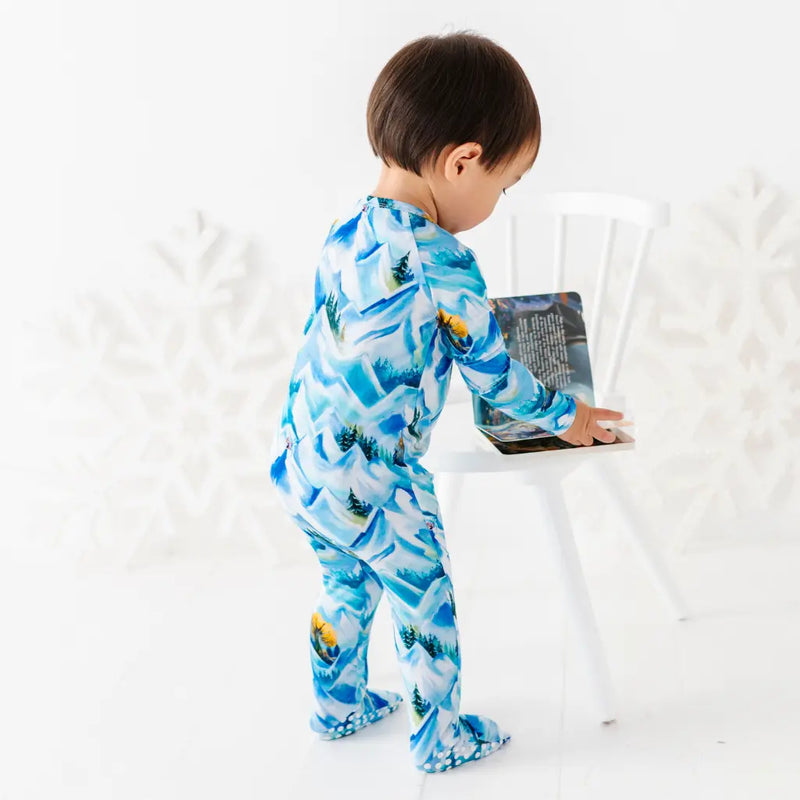 Child wearing a blue and white patterned onesie standing next to a white chair.