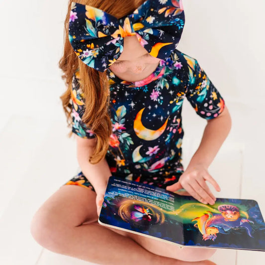 Child wearing a colorful outfit with a large bow, holding a book with a similar design.