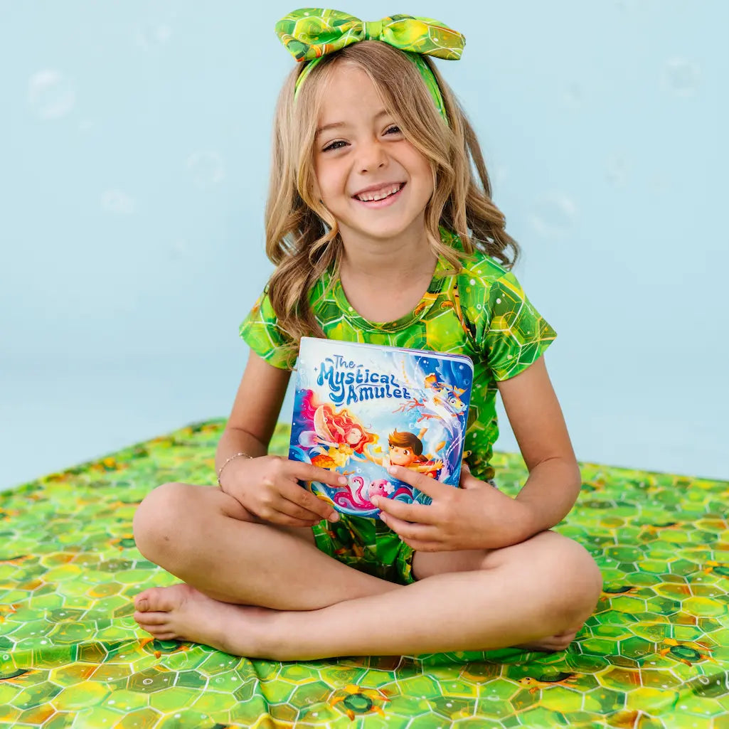 Young girl in green outfit with bow holding a book, sitting on a green and yellow textured surface.