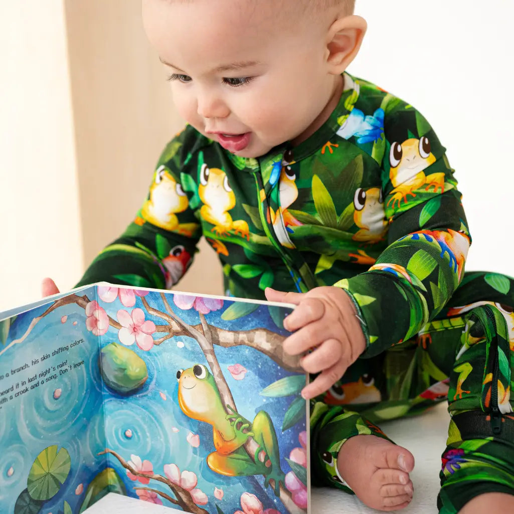 Baby in a colorful onesie reading a book with frog illustrations.