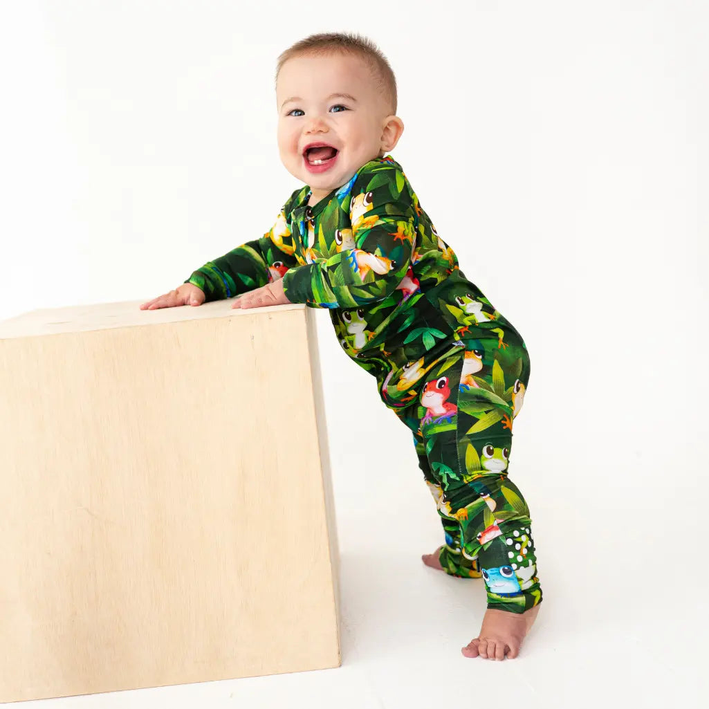 Baby smiling and standing next to a wooden block, wearing a dark green jungle print onesie on a white background