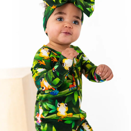 Baby wearing a dark green frog print outfit with matching headband on a white background