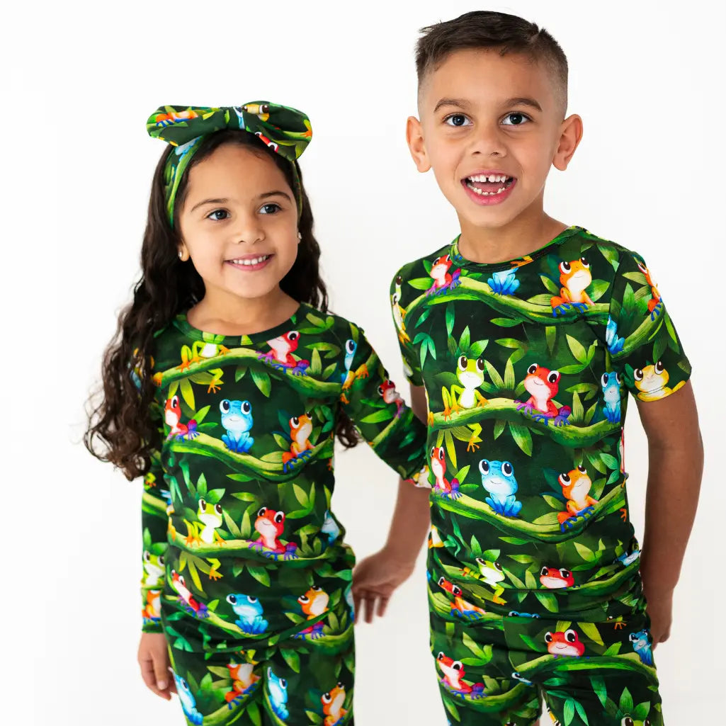 Two children wearing matching green outfits with colorful frog patterns on a white background