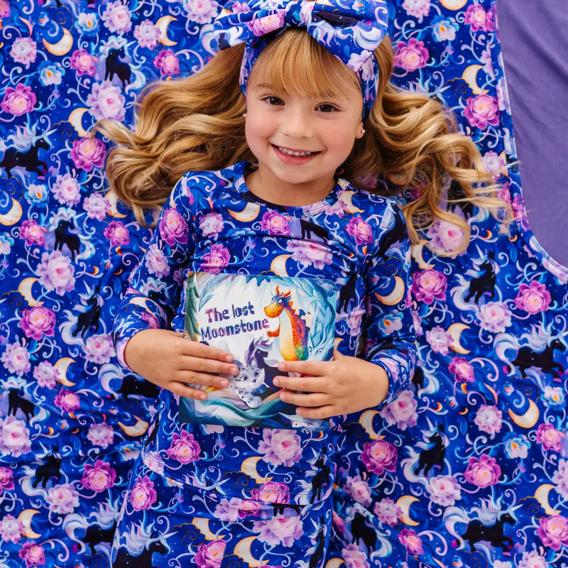 Child in a blue unicorn-themed outfit holding a book, lying on a matching blanket.