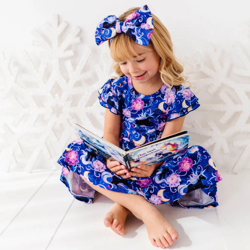 Child wearing a blue floral dress and headband, sitting on a white surface with a book.