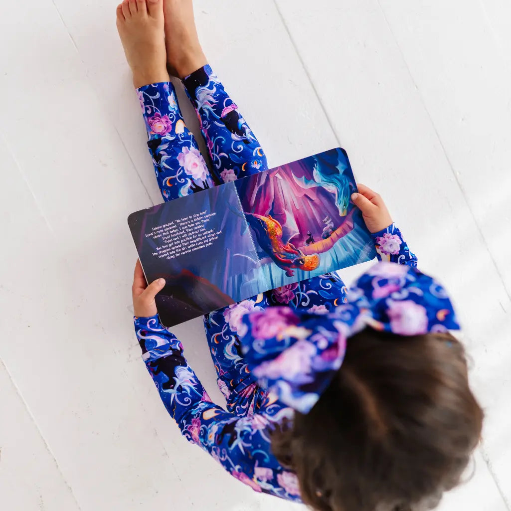 Child wearing colorful leggings and headband, holding a book with a mermaid illustration on a white floor.