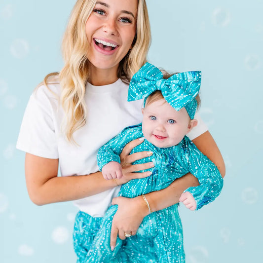 Woman holding a baby both wearing matching turquoise outfits with large bows against a light blue background