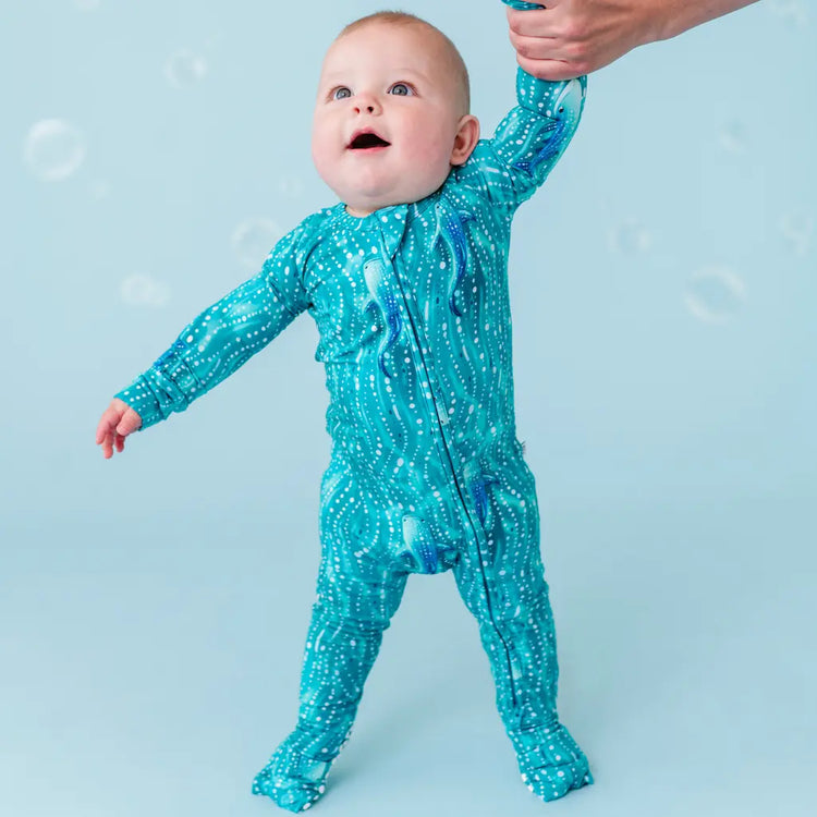A baby wearing a teal blue one-piece zipper pajama with a whale shark and shimmering teal waves pattern, standing with the help of an adult hand.