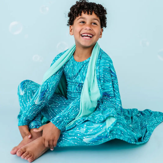A child sitting on the floor smiling while wrapped in a blue blanket with a whale shark and underwater pattern.