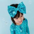 A child with dark hair styled with a large bow headband featuring a teal color with white spots and shimmering teal waves pattern, matching the child's top.