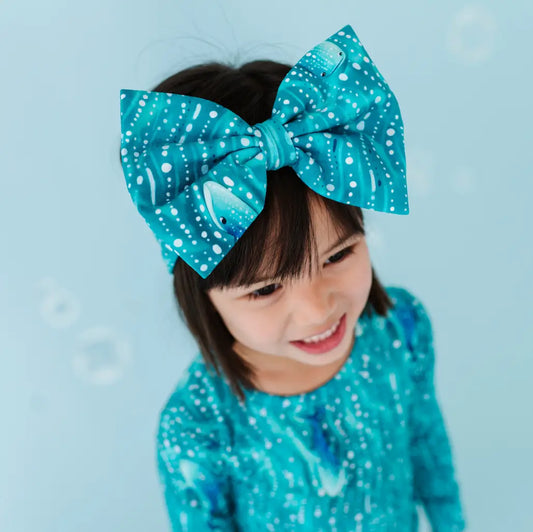 A child with dark hair styled with a large bow headband featuring a teal color with white spots and shimmering teal waves pattern, matching the child's top.