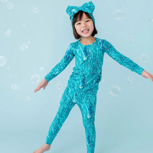 A child wearing a teal long sleeve top and pants with a whale shark and ocean wave pattern, along with a matching headpiece, against a blue background with white dots.