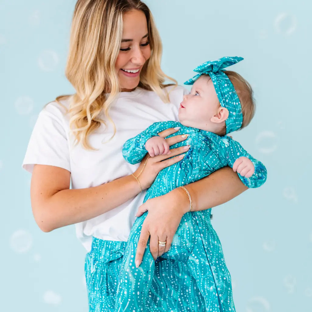 Woman holding a baby dressed in teal outfits against a light blue background.