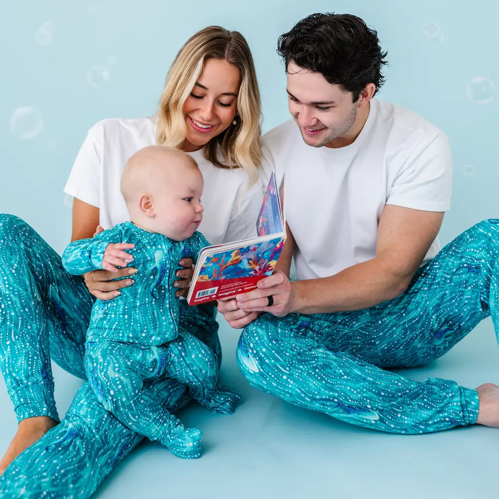 A family sitting on a blue background, with the father and mother reading a book to their young child who is wearing matching blue pajama pants with a whale print.