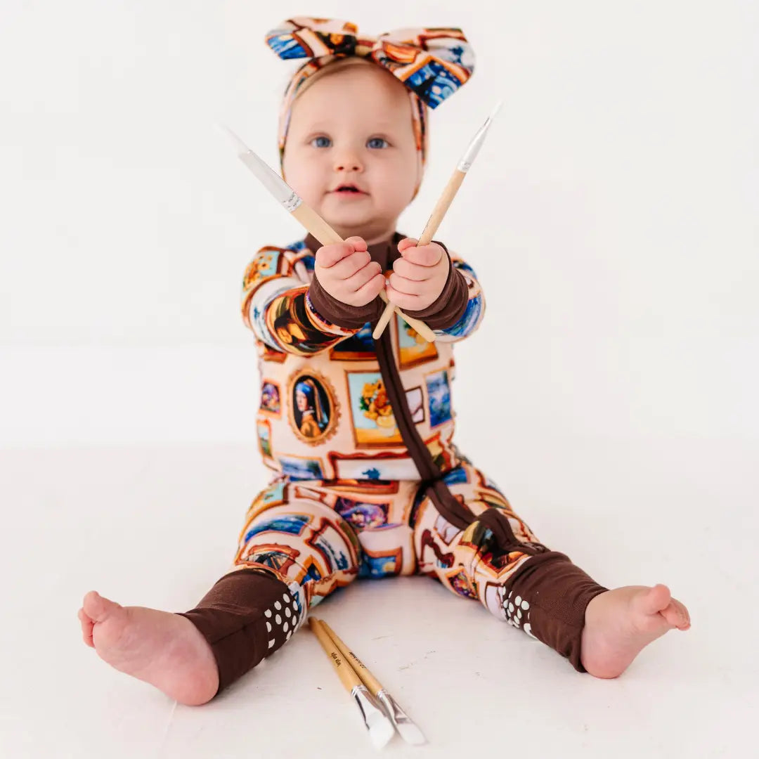A baby sitting on the floor holding paintbrushes, wearing a framed-paintings print zip-up pajama and matching bow headband.