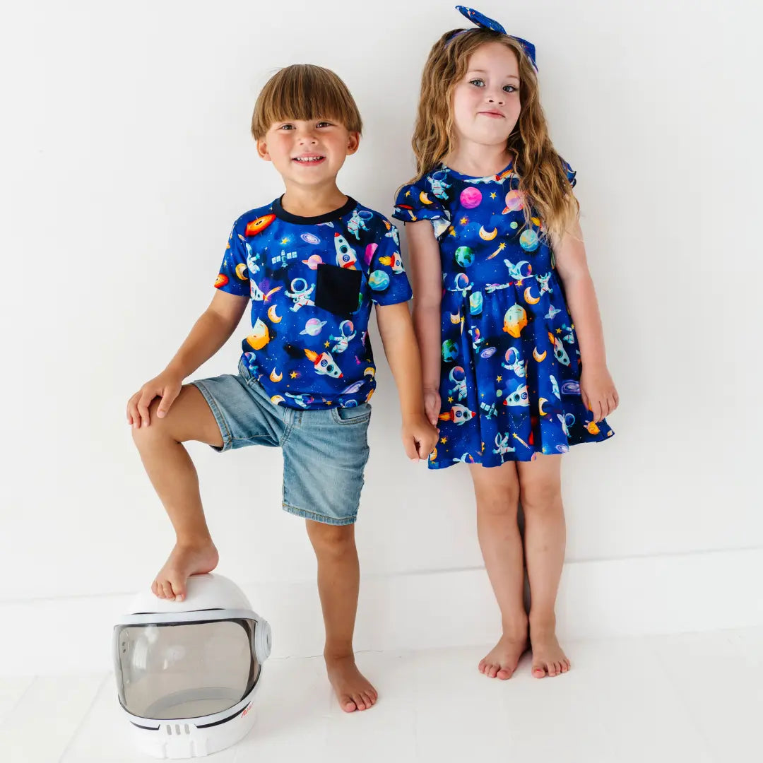 A boy and a girl standing against a white wall wearing matching navy space-print pajamas, one a tee and shorts and one a flutter sleeve dress, with a toy astronaut helmet on the floor.