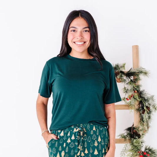 A woman with a big smile in a solid dark green short sleeve tee, shown from the waist up with jogger pajama pants in a dark green print of gold Christmas trees.