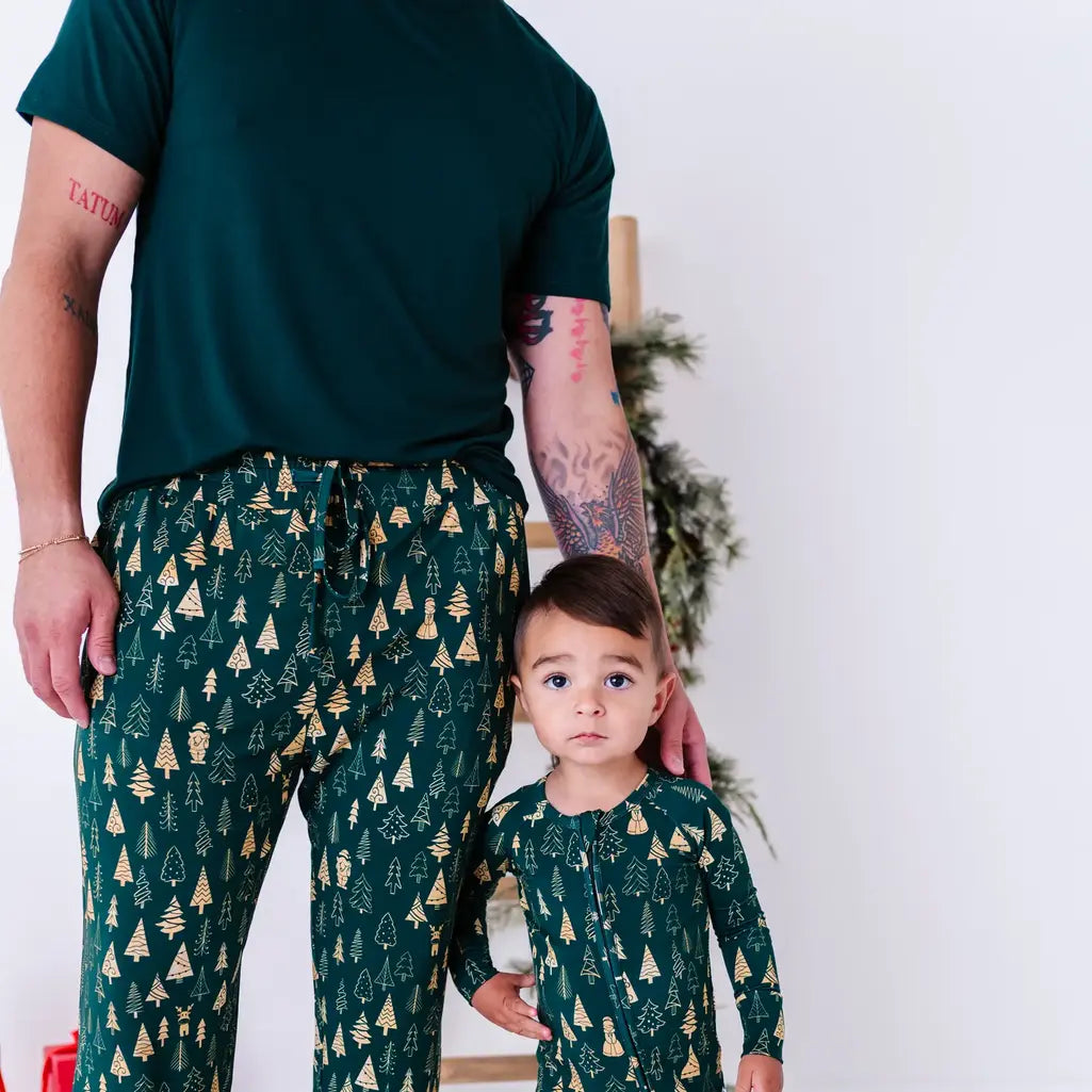 An adult in a dark green tee and pajama pants standing beside a toddler in a zip up pajama, both in a dark green print of gold Christmas trees.