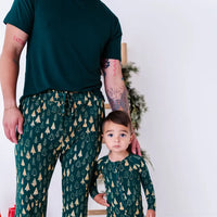 An adult in a dark green tee and pajama pants standing beside a toddler in a zip up pajama, both in a dark green print of gold Christmas trees.