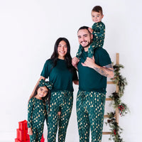 A family of four smiling at the camera with a child on an adult's shoulders, in matching pajamas in a dark green print of gold Christmas trees and dark green tees.