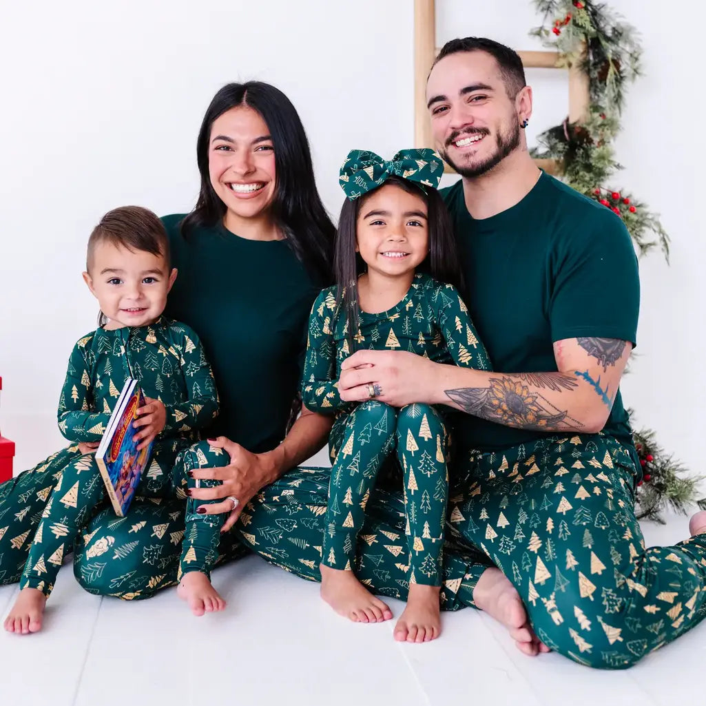 A family of four sitting close together and smiling at the camera in matching pajamas in a dark green print of gold Christmas trees and dark green tees.