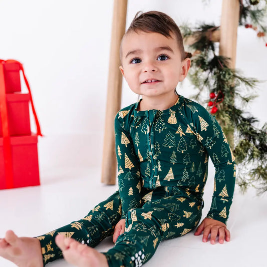 A toddler smiling at the camera while sitting barefoot on a white floor, in a zip up pajama in a dark green print of gold Christmas trees.