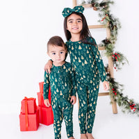 An older sibling with an arm around a younger one, both smiling in a pajama set and zip up baby pajama in a dark green print of gold Christmas trees.