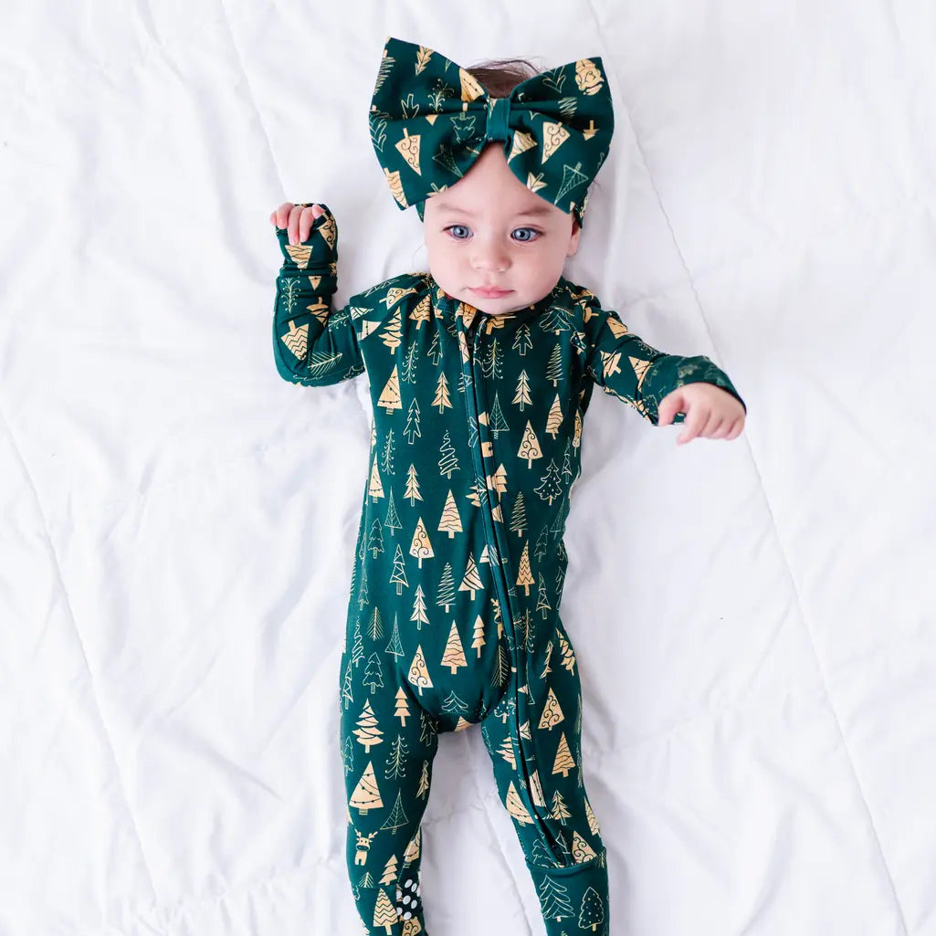 A baby in a bow headband lying on a white quilt with one hand raised, in a zip up footed pajama in a dark green print of gold Christmas trees.
