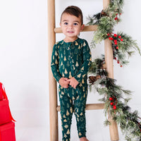 A smiling toddler standing with hands clasped in front of a wooden ladder with a garland, in a zip up pajama in a dark green print of gold Christmas trees.