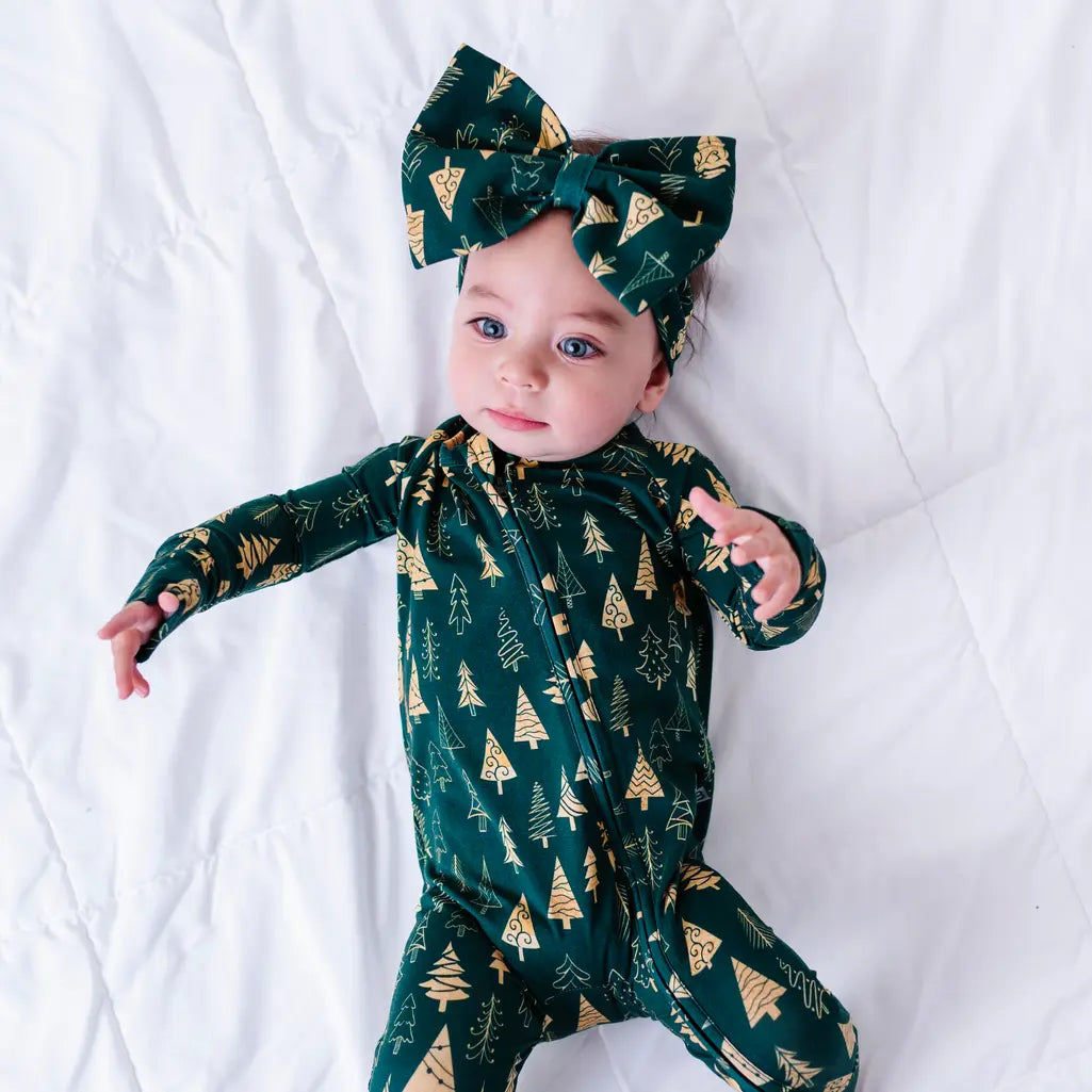 A baby in a bow headband looking at the camera while lying on a white quilt with arms stretched out, in a zip up pajama in a dark green print of gold Christmas trees.