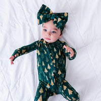 A baby in a bow headband looking at the camera while lying on a white quilt with arms stretched out, in a zip up pajama in a dark green print of gold Christmas trees.