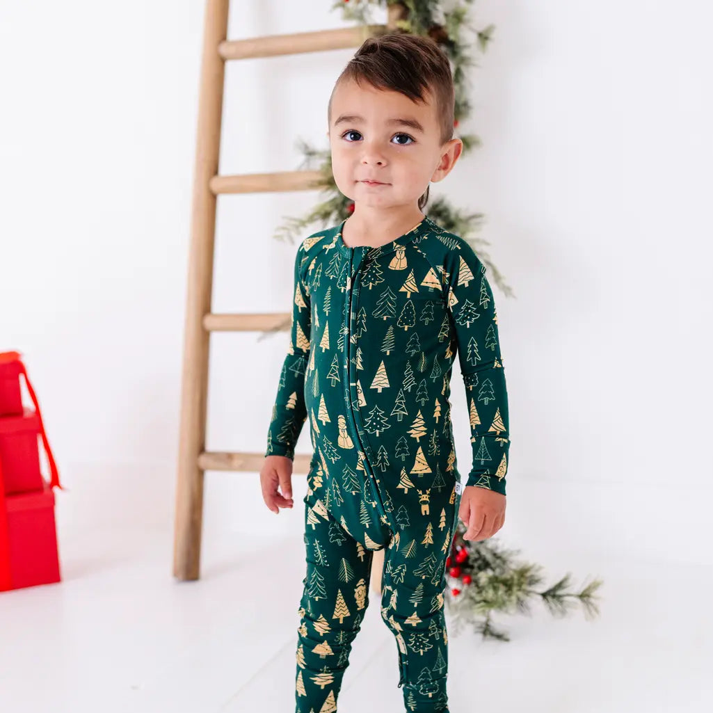 A toddler turned slightly to one side and looking at the camera in front of a wooden ladder, in a zip up pajama in a dark green print of gold Christmas trees.