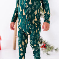 Close up of the zipper front and legs of a zip up pajama on a standing toddler, in a dark green print of gold Christmas trees.