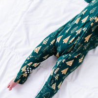 Close up of the legs of a baby lying on a white quilt, showing the folded foot cuff and grip dots of a zip up pajama in a dark green print of gold Christmas trees.