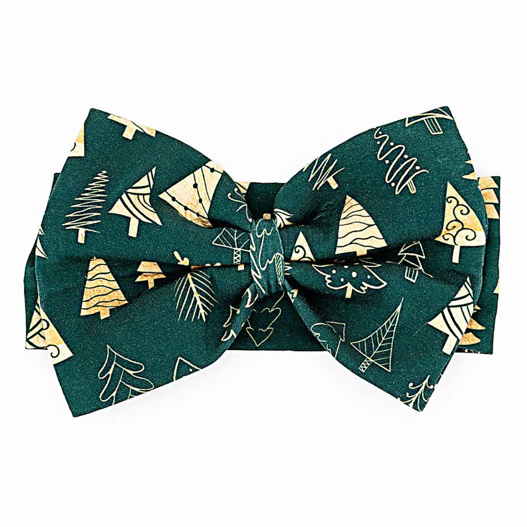 Flat lay of a large fabric bow headband in a dark green print of gold and outlined Christmas trees on a white background.