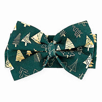 Flat lay of a large fabric bow headband in a dark green print of gold and outlined Christmas trees on a white background.