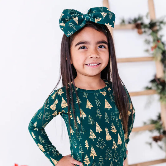 A smiling child with long dark hair and hands on hips, wearing a large fabric bow headband in a dark green print of gold and outlined Christmas trees, with a matching long sleeve pajama top.