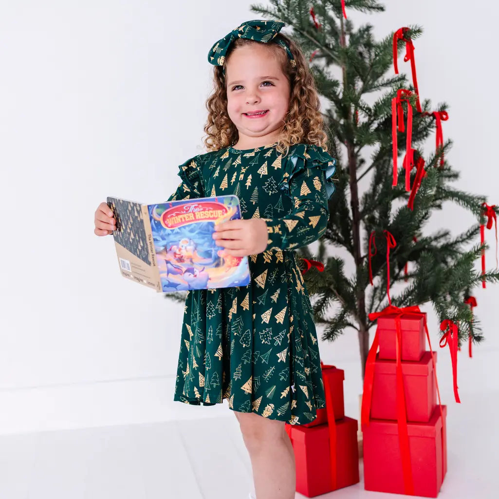 A child smiling at the camera while holding an open storybook, standing in a long sleeve twirl dress in a dark green print of gold Christmas trees.