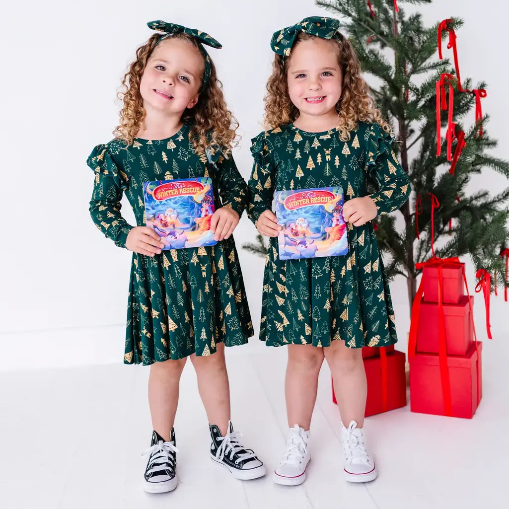 Two kids holding storybooks and smiling in matching long sleeve twirl dresses in a dark green print of gold Christmas trees.