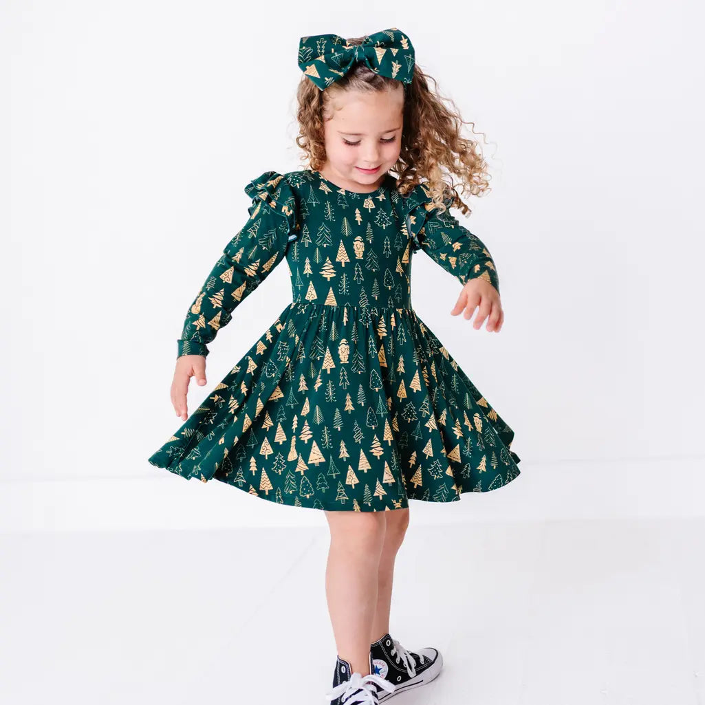 A child twirling with the full skirt flared wide, in a long sleeve twirl dress with flutter ruffles at the shoulders in a dark green print of gold Christmas trees.