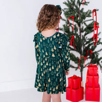 Back view of a child in a long sleeve twirl dress with flutter ruffles at the shoulders in a dark green print of gold Christmas trees.