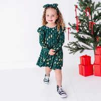 A child walking toward the camera with a smile, in a long sleeve twirl dress with flutter ruffles at the shoulders in a dark green print of gold Christmas trees.
