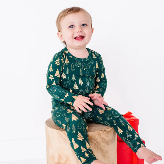 A toddler laughing and sitting barefoot on a wooden stump, in a long sleeve pajama top and matching pants in a dark green print of gold Christmas trees.