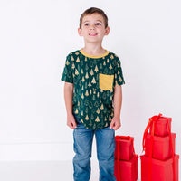 A child standing in jeans and looking up with a calm smile, in a short sleeve pocket tee with a mustard ribbed collar and pocket in a dark green print of gold Christmas trees.
