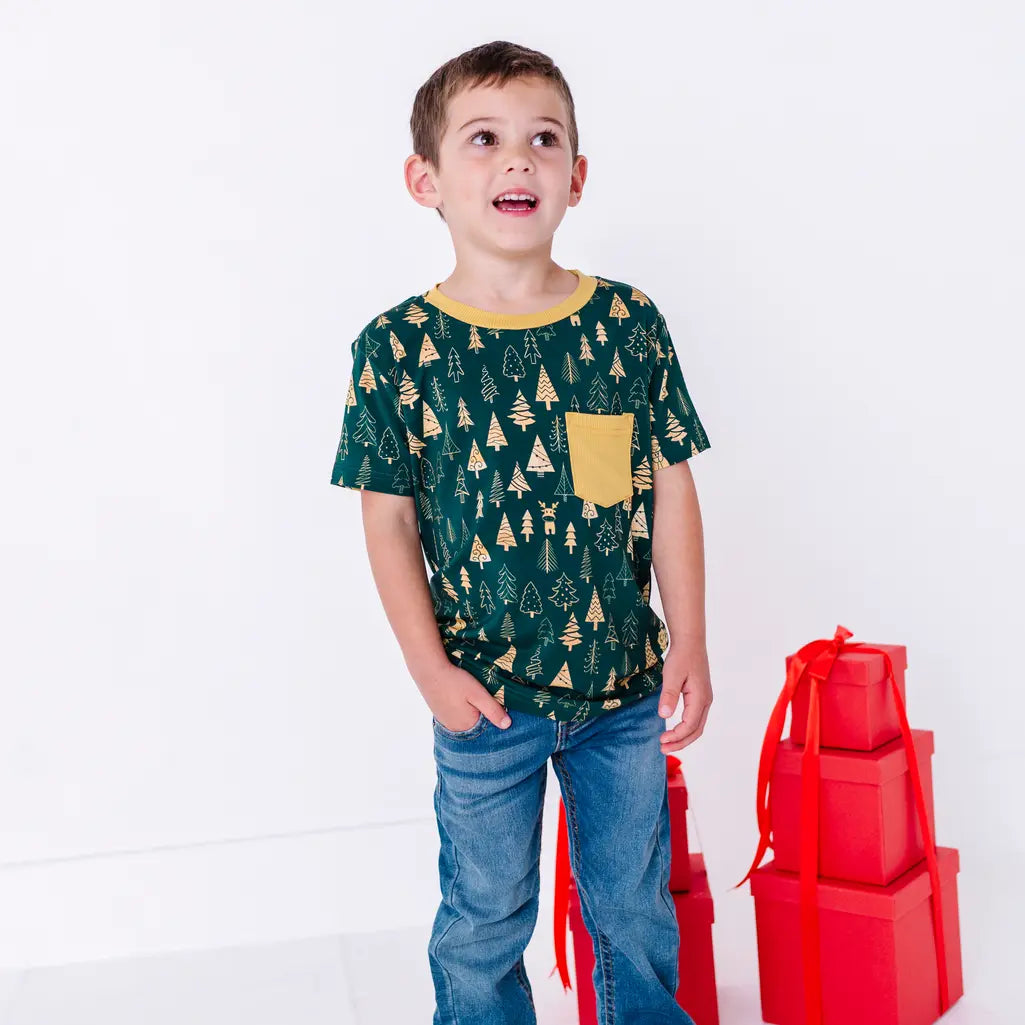 A child looking up with an open smile and hands in the jeans pockets, in a short sleeve pocket tee with a mustard pocket in a dark green print of gold Christmas trees.